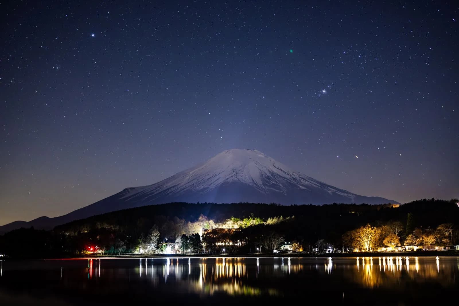 Mount fuji at night with starry sky and illuminated lakeside buildings reflecting on water surface