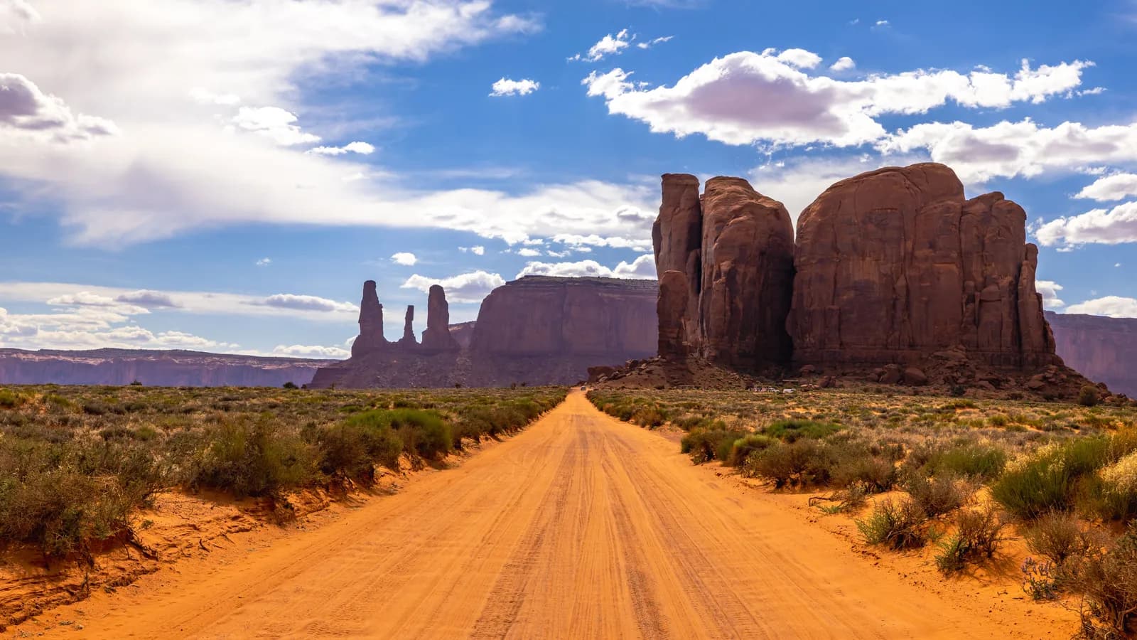 Scenic dirt road leading through monument valley desert landscape under a bright blue cloudy sky