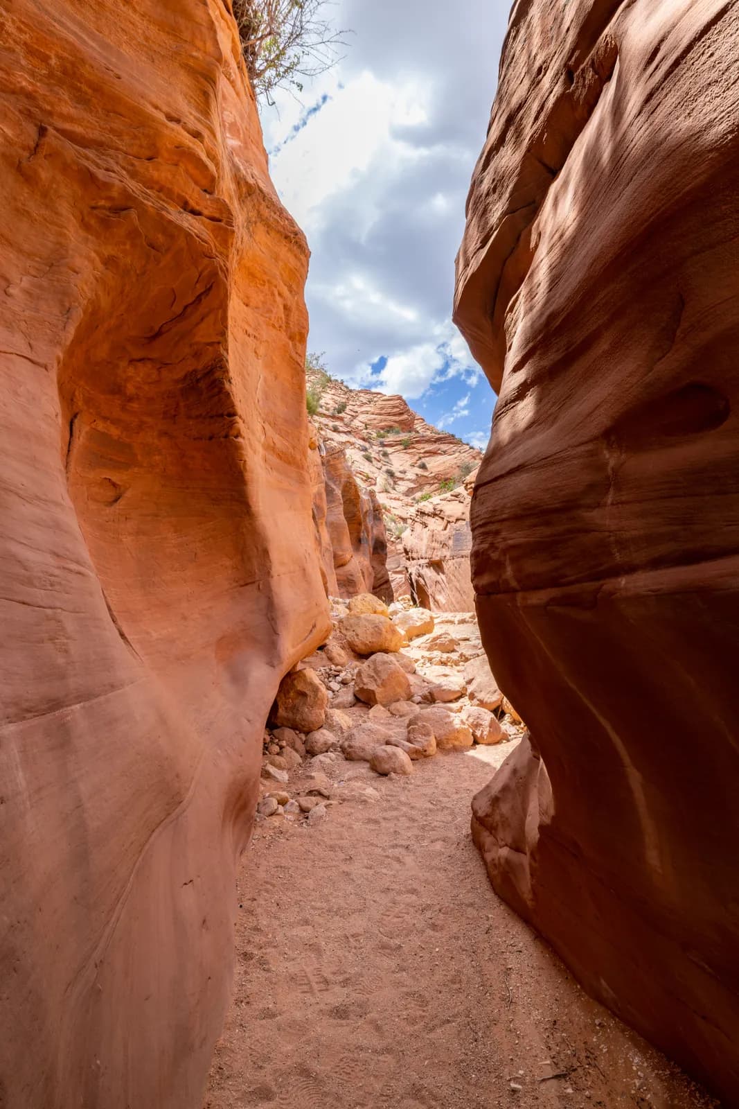 Narrow slot canyon with red sandstone walls and sandy path under a bright blue cloudy sky