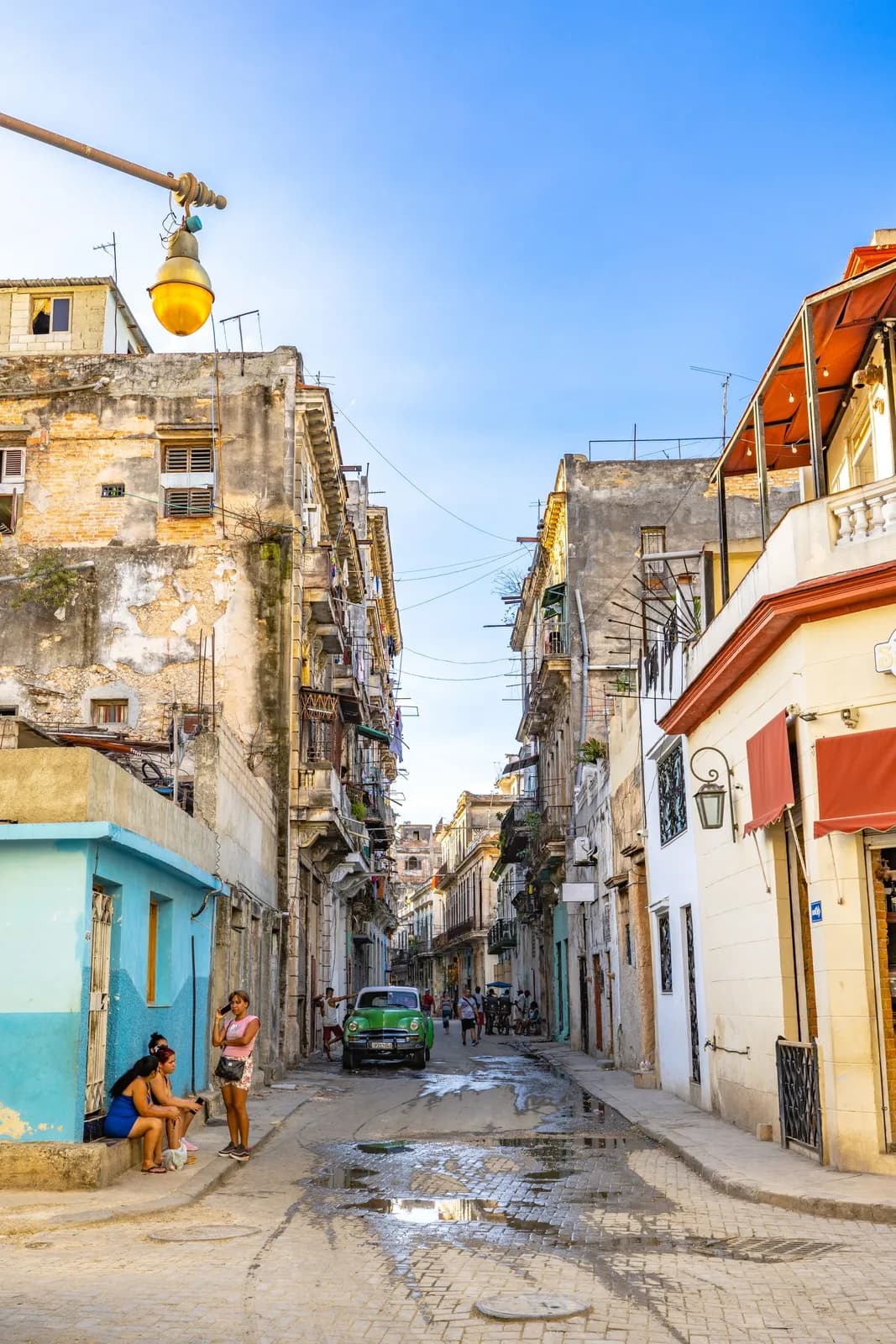 Narrow street in old havana with vintage car and people sitting on sidewalk in cuba