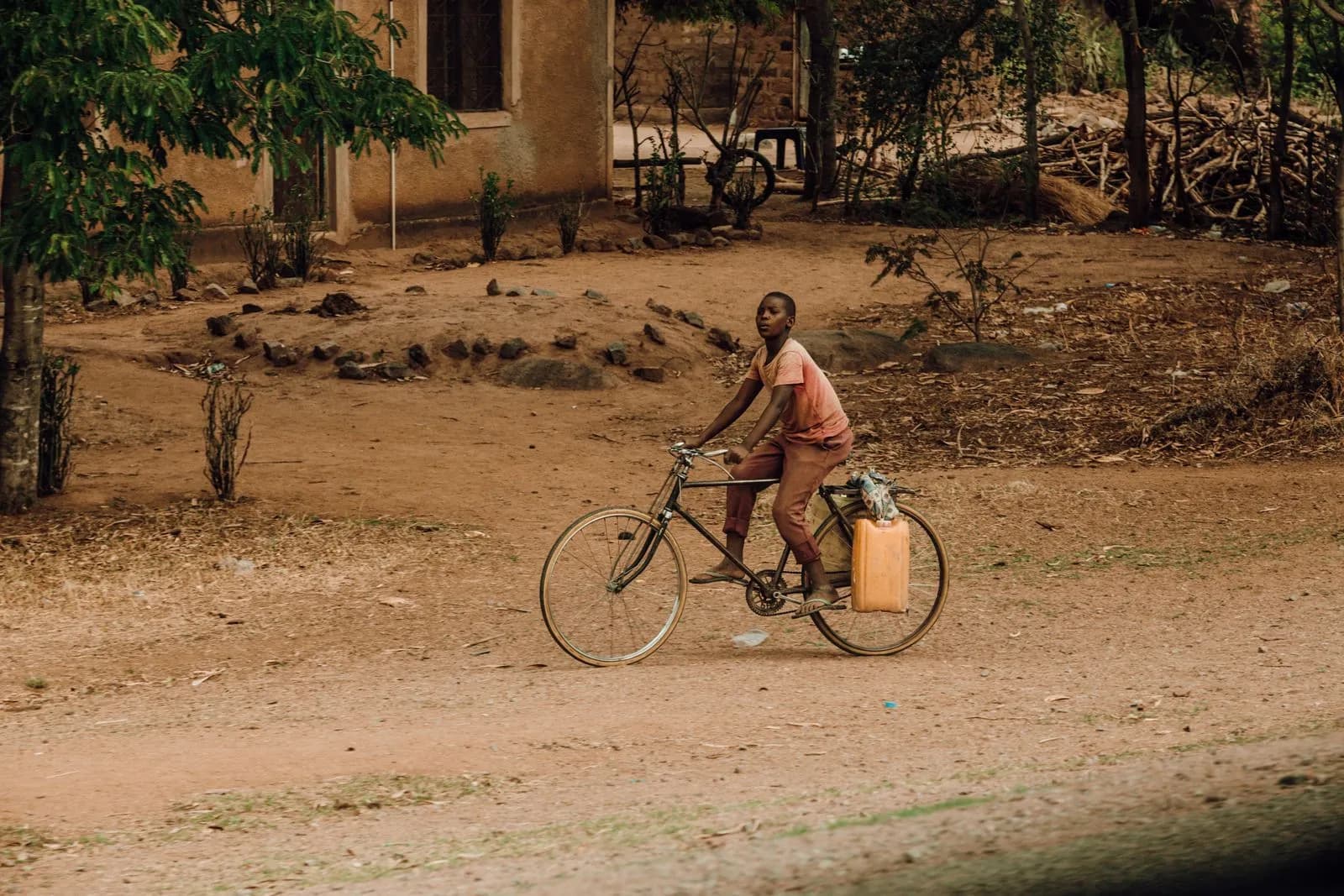 Young boy riding a bicycle on a dirt road carrying a plastic water container in a rural village