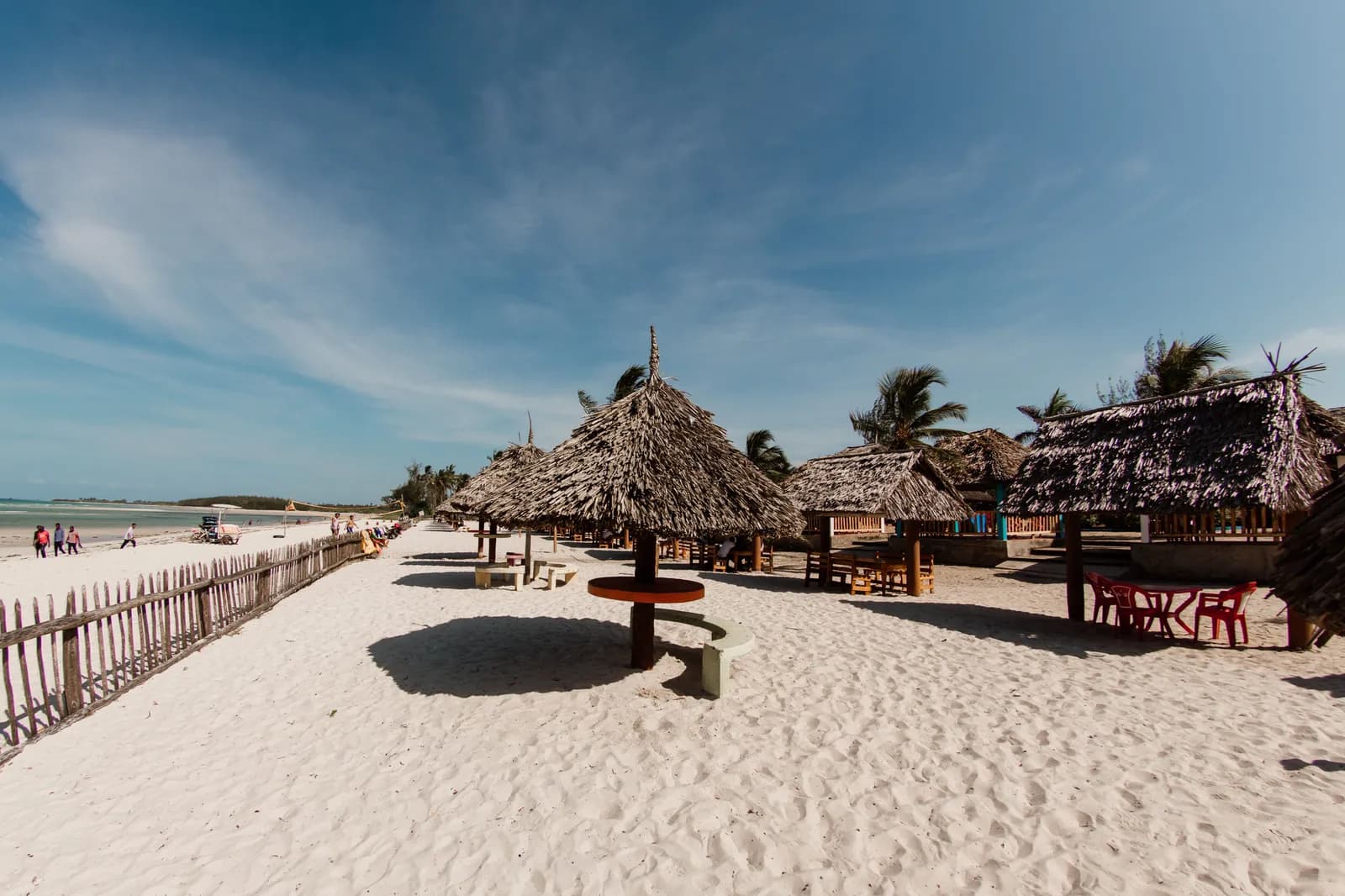 Tropical beach resort with thatched roof umbrellas and wooden tables on white sand under blue sky