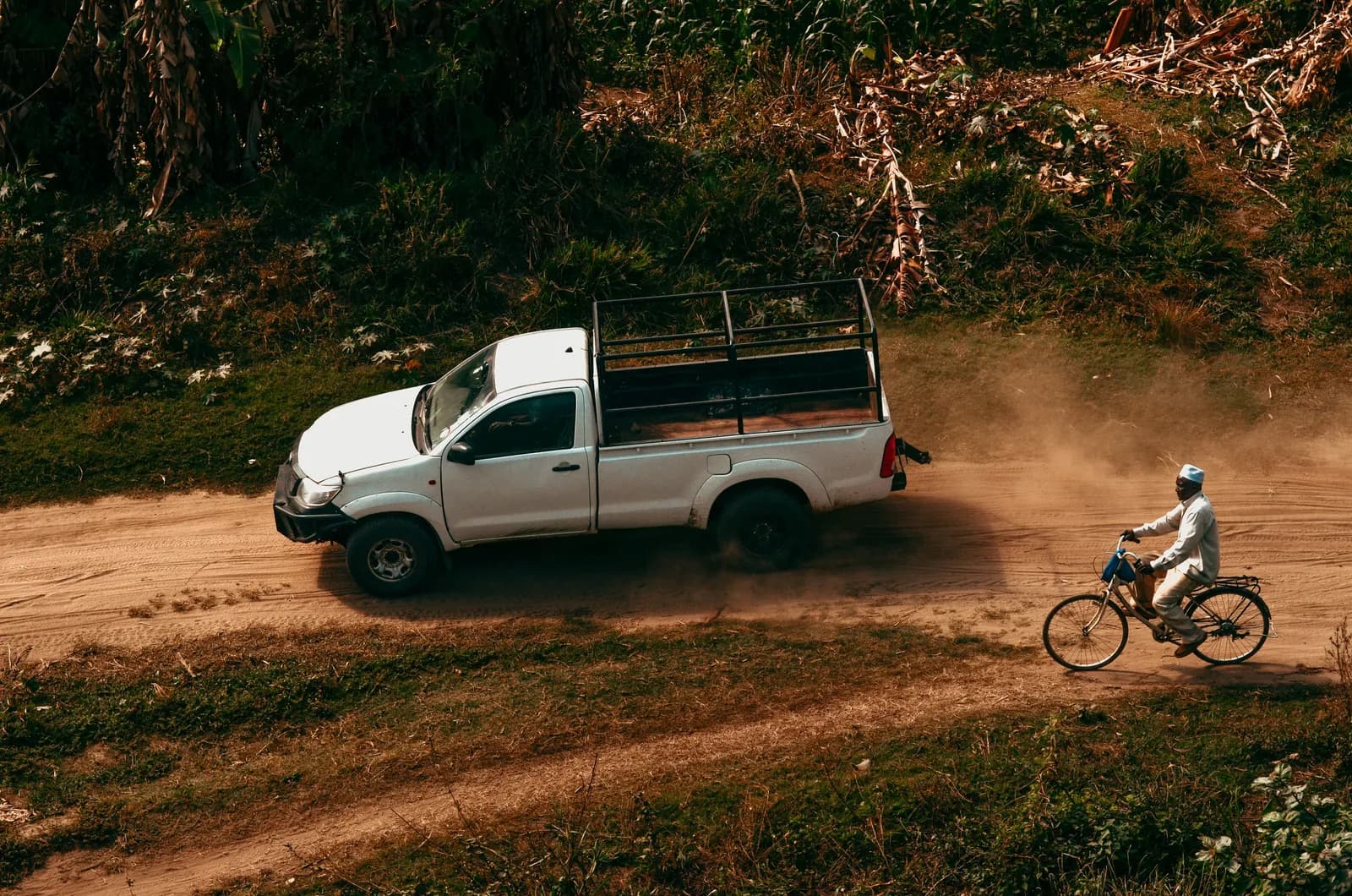 Man riding bicycle alongside white pickup truck on dirt road through forest