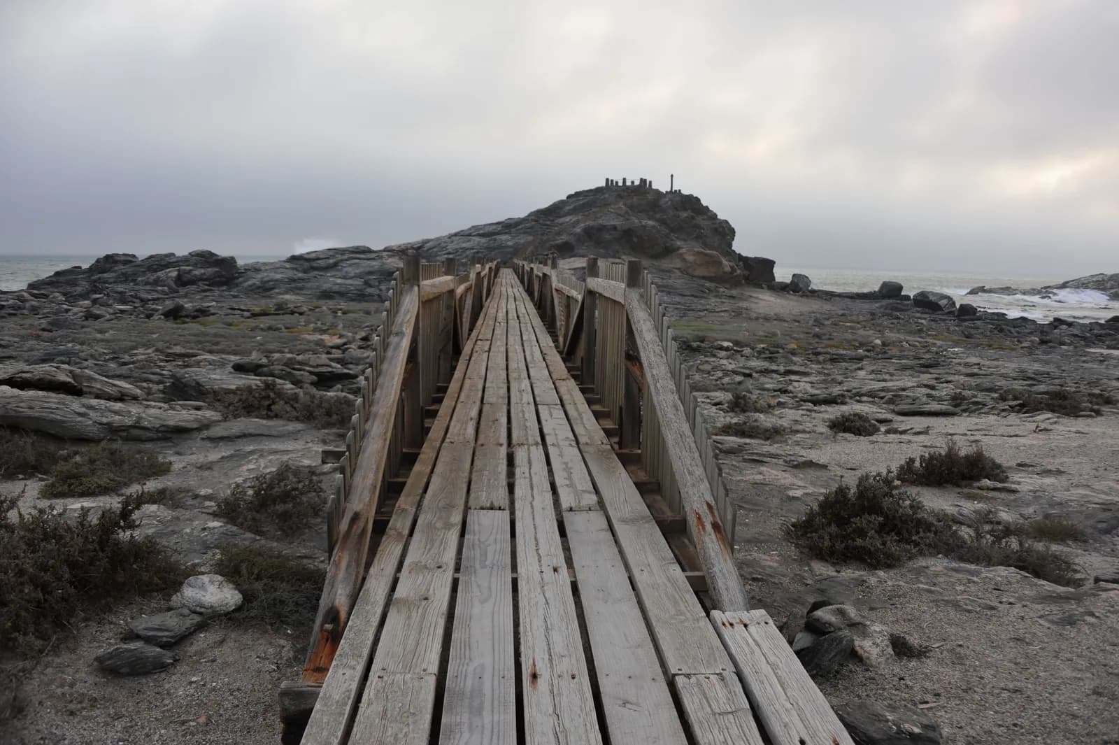Wooden footbridge leading to a rocky island on a cloudy day at the ocean coast