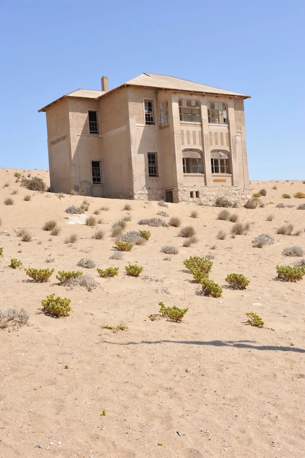 Abandoned house in the desert landscape with sand dunes and dry vegetation under clear blue sky