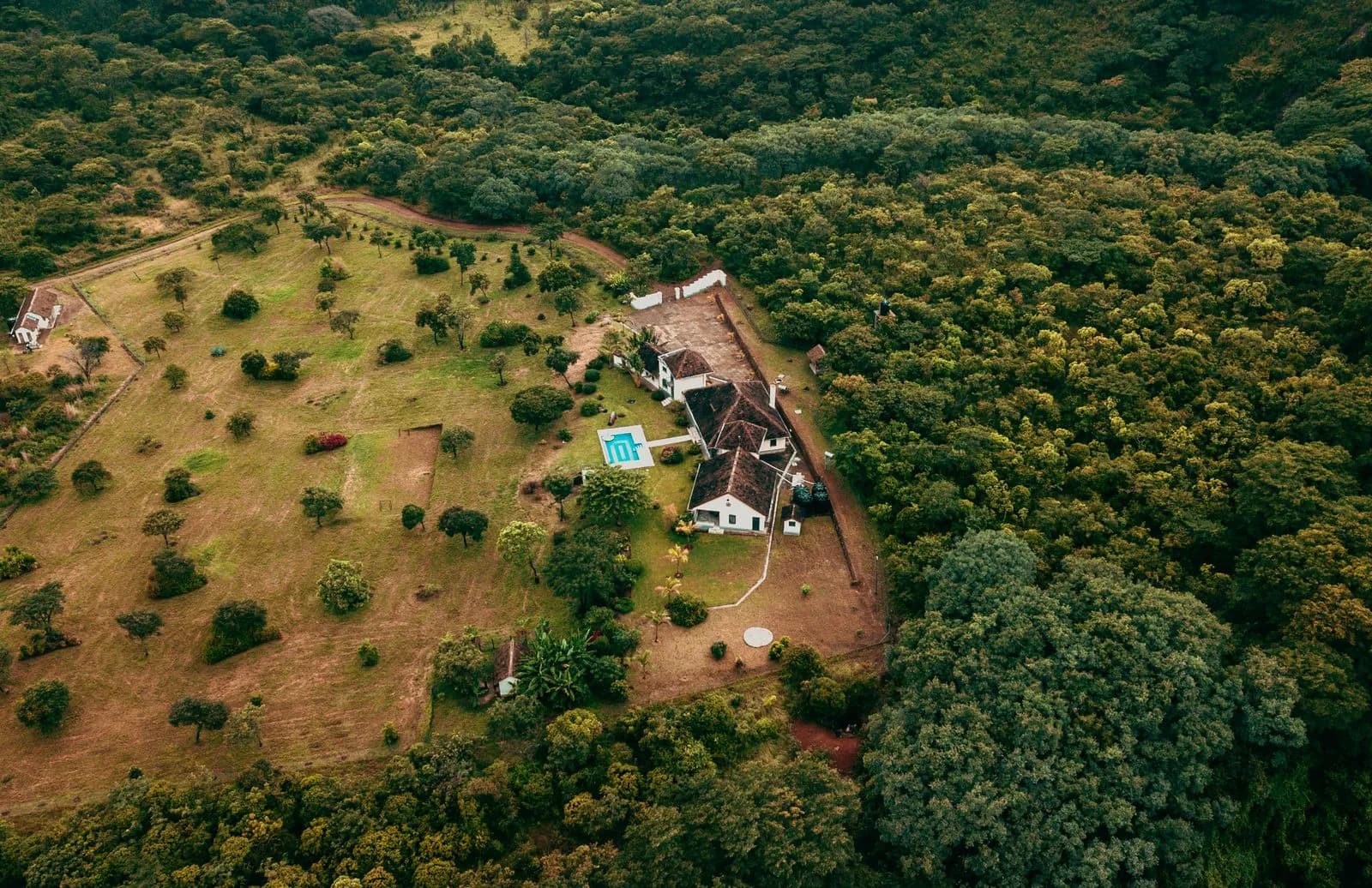 Aerial view of a rural farmhouse with a swimming pool surrounded by lush green forest landscape