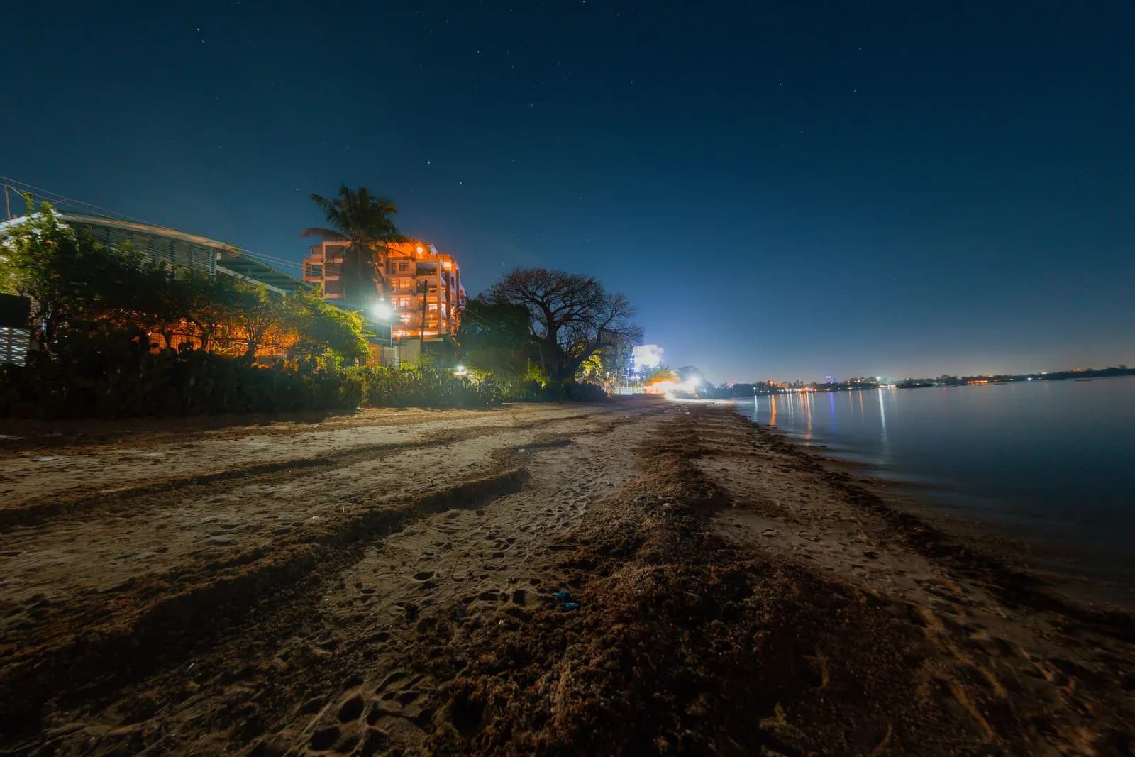Night view of a sandy beach with illuminated coastal buildings and calm ocean water at twilight