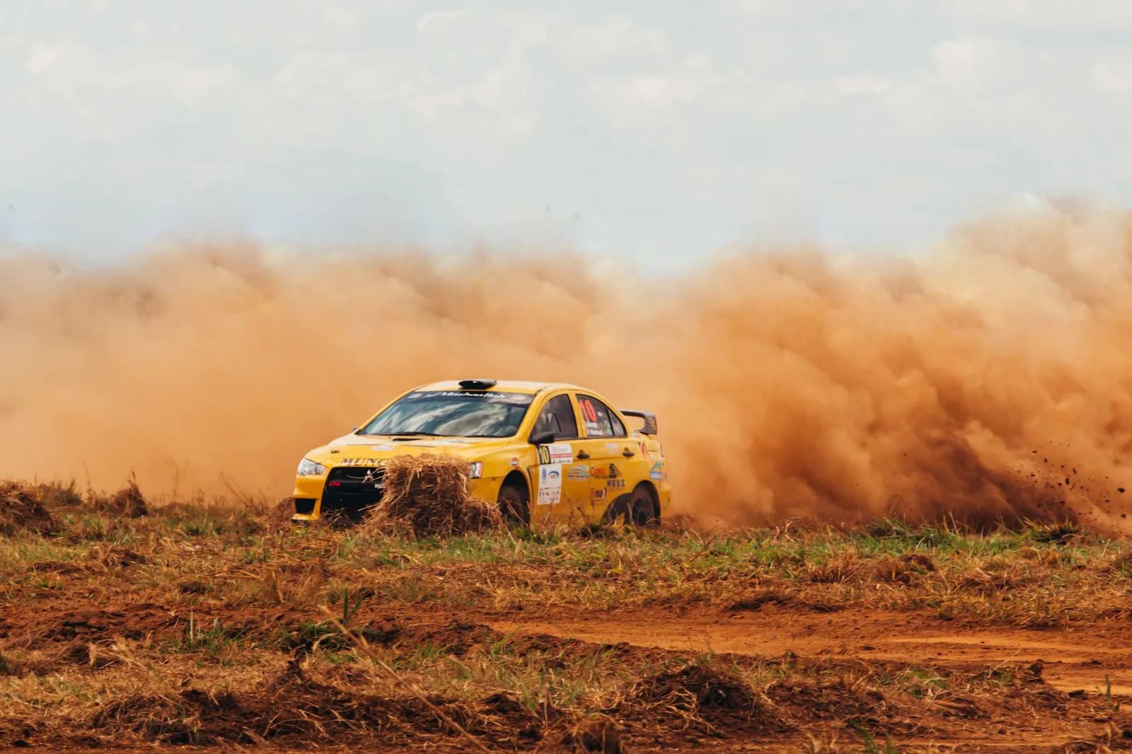Yellow rally car driving fast on dirt track creating large dust cloud during professional race event