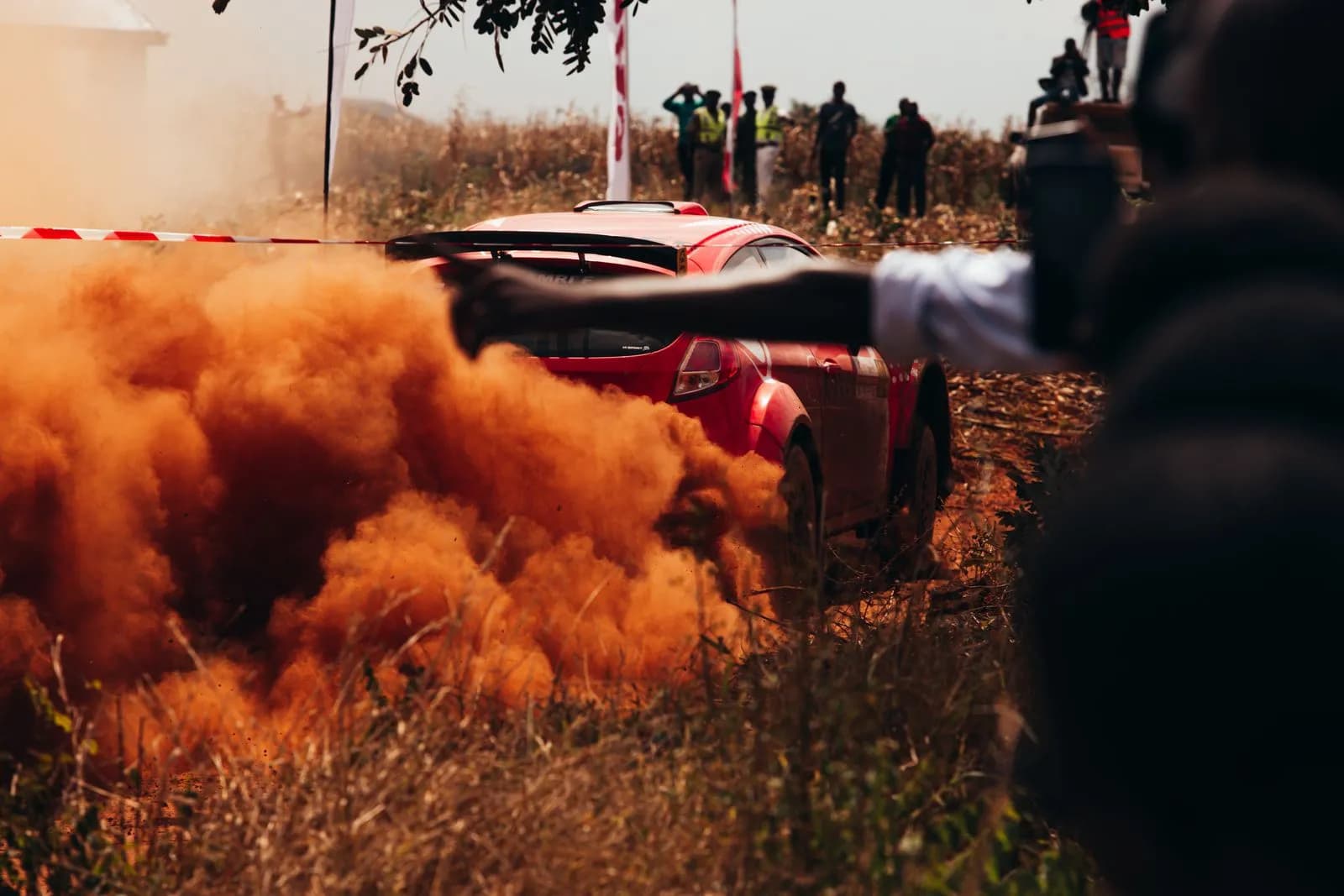 Red rally car driving fast on dirt track with orange dust cloud and spectators watching the race