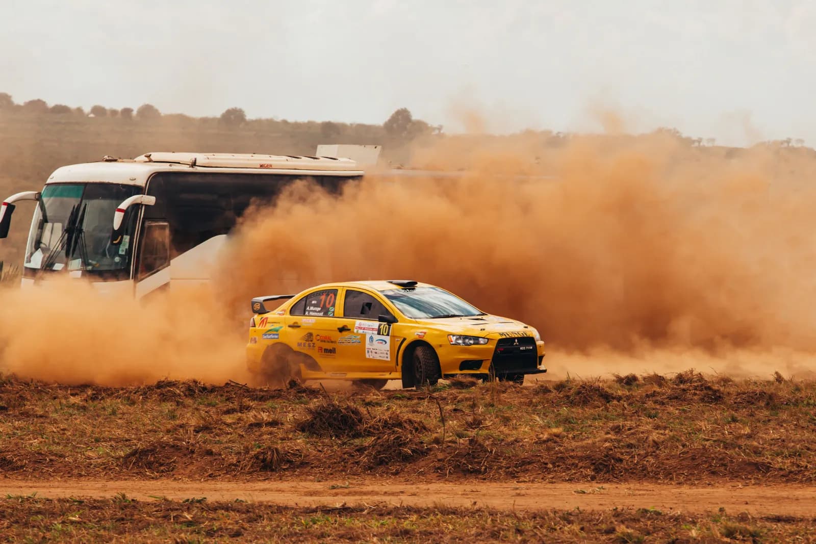 Yellow rally car driving fast on dirt track with dust clouds and bus in background