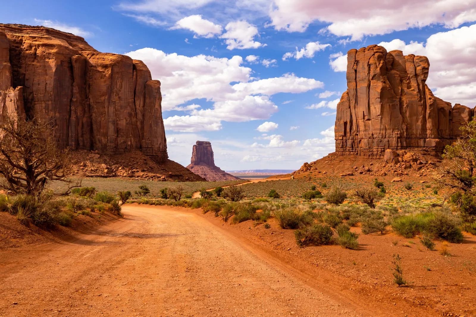 Scenic dirt road leading through monument valley desert landscape under blue sky with clouds