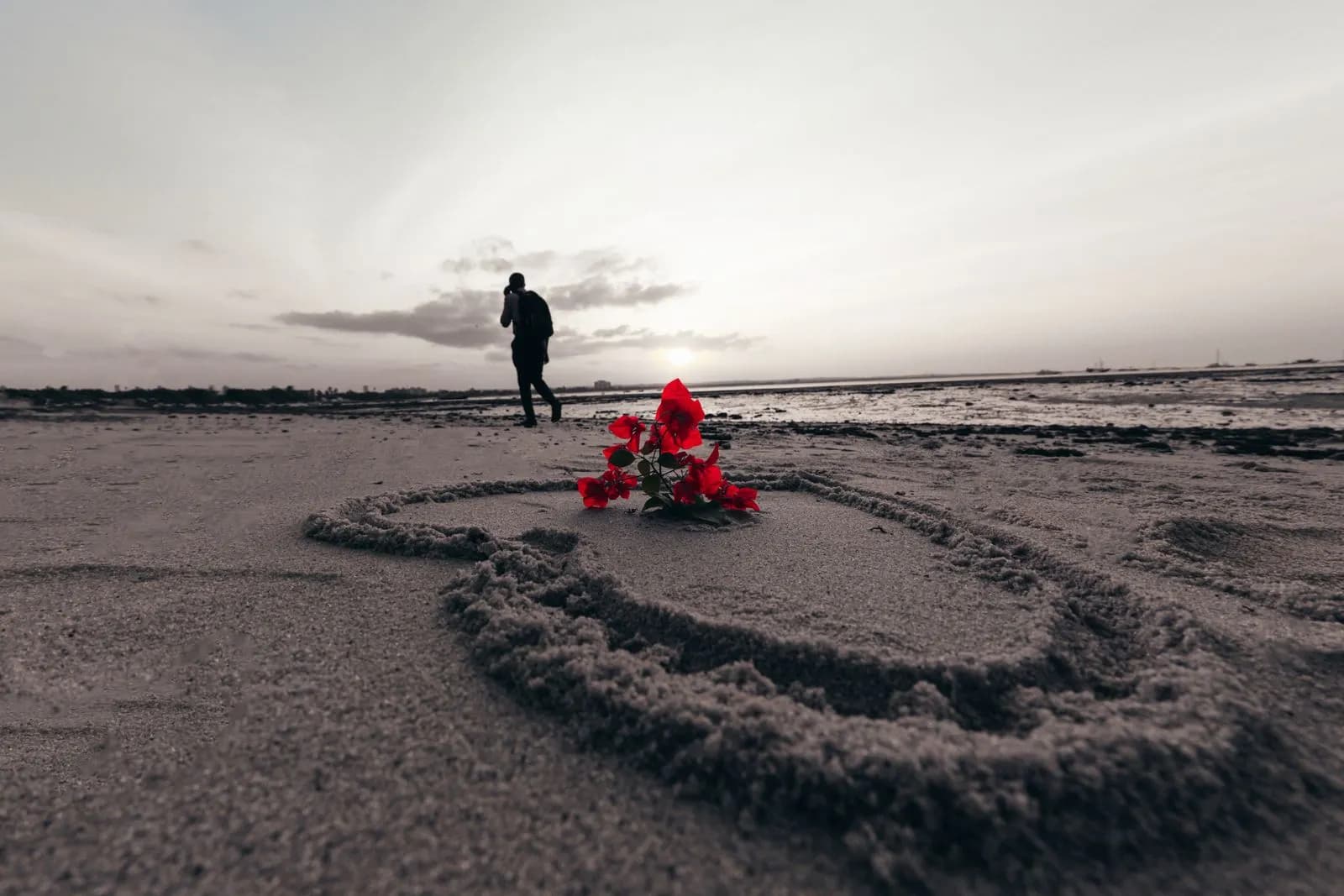Red flower inside a heart shape drawn on the beach sand with a man walking in the distance